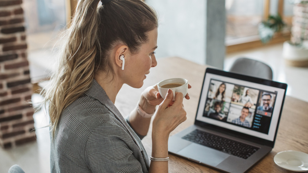 Frau sitzt mit einem Kaffee vor einem Laptop auf dem ein Online-Meeting stattfindet