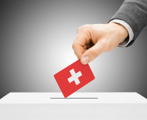 Männliche Hand legt roten Wahlzettel mit Schweizerkreuz in Wahlurne